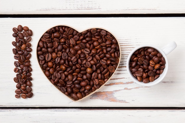 Ik hou van natuurlijk vers koffieconcept. Koffiezaden gerangschikt in een hartvorm en vullende beker. Wit houten oppervlak.