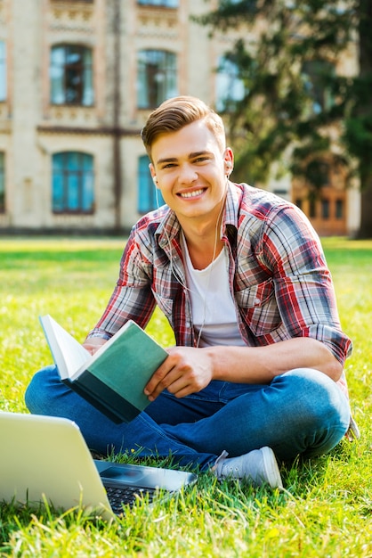 Ik hou van studeren. Vrolijke mannelijke student die boek leest en glimlacht terwijl hij op het gras zit en voor het universiteitsgebouw