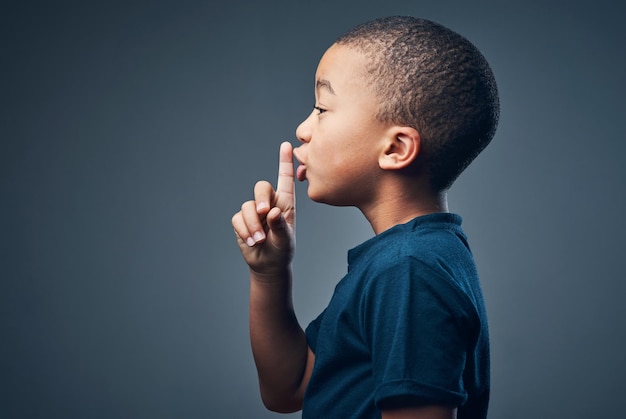 Ik kan een geheim bewaren als je kunt studio-opname van een schattige kleine jongen die poseert met zijn vinger op zijn lippen tegen een grijze achtergrond