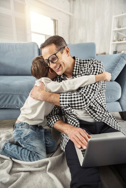 Ik koester jou. Knappe uitbundige donkerharige vader zittend op de vloer en bezig met zijn laptop en zijn zoon hem knuffelen