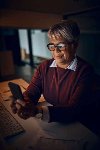 Ik kom over een paar minuten naar huis Shot van een volwassen zakenvrouw die een mobiele telefoon gebruikt terwijl ze laat op kantoor aan het werk is