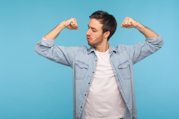 Ik kreeg macht Portret van een knappe man in een denim overhemd van een werknemer die handen opsteekt en met verrukking naar biceps kijkt, zelfverzekerd sterk en succesvol indoor studio-opname geïsoleerd op blauwe achtergrond