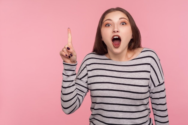 Ik weet het antwoord. Portret van een geïnspireerde vrouw in een gestreept sweatshirt die met de vinger omhoog wijst en een goed idee heeft, verbaasd verrast door een plotseling uitgevonden slimme oplossing. indoor studio-opname geïsoleerd