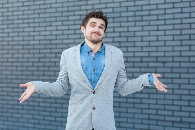 Ik weet het niet. Portret van een verwarde knappe bebaarde man in een ongedwongen stijl, staande, opgeheven armen en kijkend naar de camera met een twijfelachtig gezicht. indoor studio opname op bakstenen muur achtergrond.