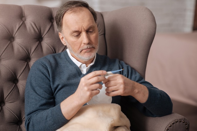 Ik werd ziek. Senior man zittend op de bank, thermometer kijken en temperatuur controleren.