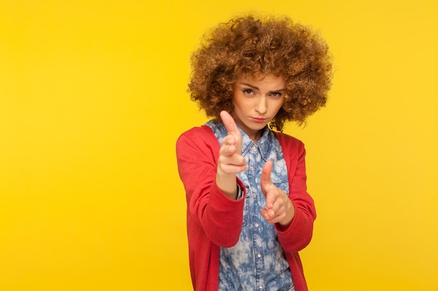 Ik zal je vermoorden. Portret van een hipstervrouw met krullend haar die vingergeweren naar de camera wijst, een doelwit richt en dreigt te schieten met een pistoolhandgebaar. indoor studio-opname geïsoleerd op gele achtergrond