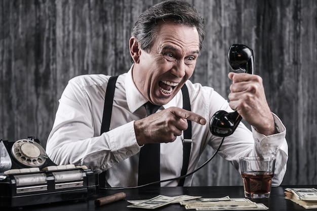 Ik zal je vinden waar je ook bent! Woedende senior man in shirt en bretels die tegen de telefoon schreeuwt terwijl hij aan tafel zit met veel geld naast hem