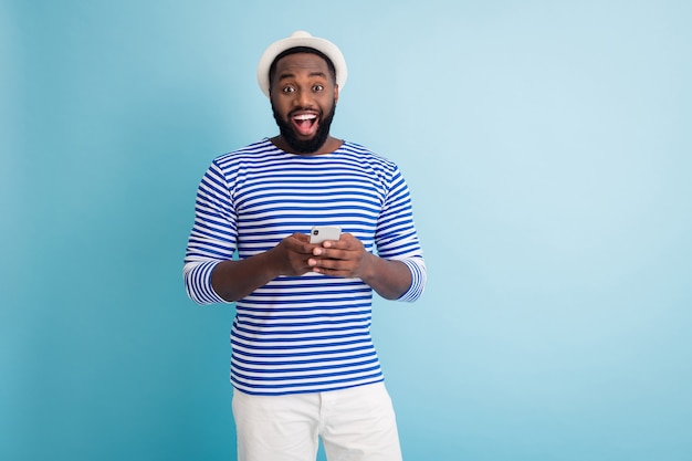 Ik zie de oceaan! Foto van grappige donkere huid man reiziger goed humeur telefoon app chatten vrienden dragen witte zonnepet gestreepte matroos hemd korte broek geïsoleerde blauwe kleur muur