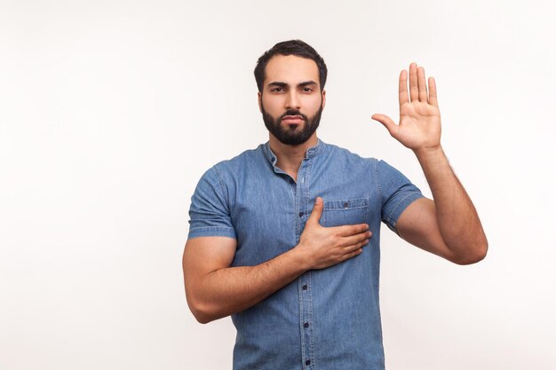 Ik zweer bewuste eerlijke man die één hand opheft en op de borst een andere vasthoudt die vloekt in eerlijkheid en toewijding patriottisme Indoor studio shot geïsoleerd op witte achtergrond