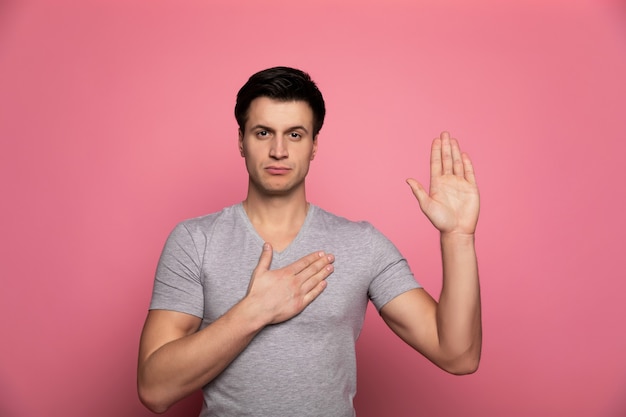 Ik zweer. serieuze man in een grijs t-shirt, die zijn rechterhand op zijn borst houdt en met zijn linkerhandpalm naar de camera kijkt.