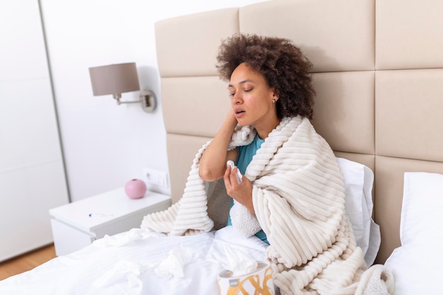 Ill african young woman covered with blanket blowing running\
nose got fever caught cold sneezing in tissue sit on sofa sick\
allergic black girl having allergy symptoms coughing at home flu\
concept