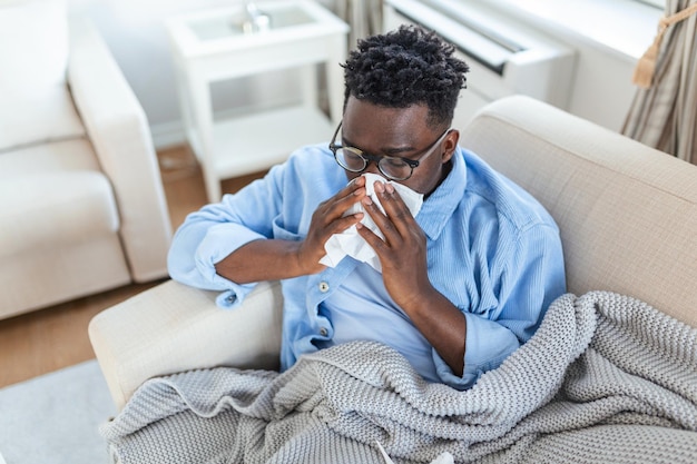 Photo ill african young woman covered with blanket blowing running nose got fever caught cold sneezing in tissue sit on sofa, sick allergic black girl having allergy symptoms coughing at home, flu conceptil