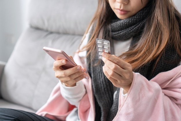 아픈 아시아 여성이 집 거실에 있는 소파에 앉아 온라인으로 알약 정보를 검색합니다.