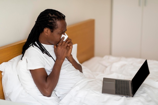 Больной черный человек сморкается и пользуется ноутбуком, сидя в постели