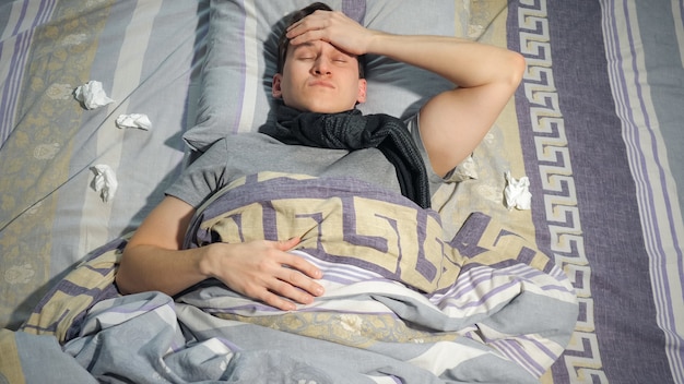 Ill guy keeping eyes closed and touching hot forehead while lying amidst used tissues on bed