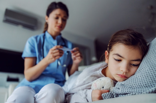 Bambina malata sdraiata sul letto a casa. in sfondo sfocato è infermiera guardando il termometro.