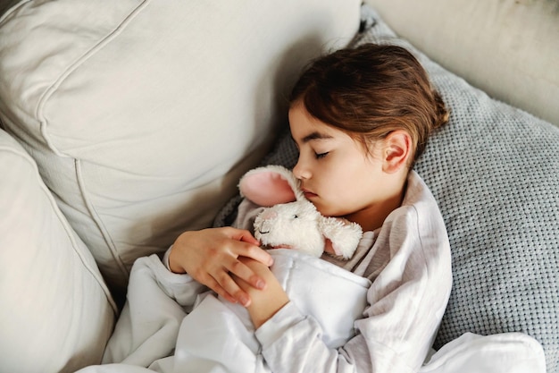 Bambina malata sdraiata sul divano di casa che abbraccia il suo coniglietto e dorme