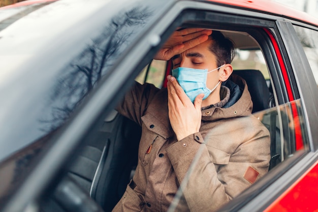 Ill man sitting in car wearing protective mask feeling sick having flu coronavirus. Prohibition driving auto with fever.