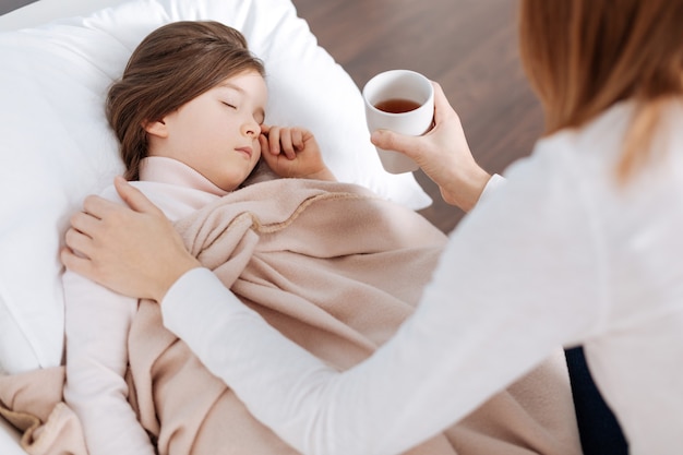 Ill pleasant little girl sleeping while her mother sitting on the sofa and holding cup of tea