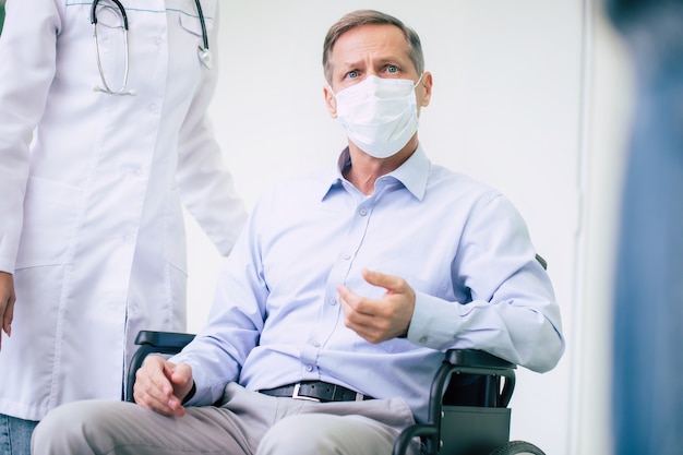 Ill senior man with protective safety mask on face in a wheelchair and a confident doctor in the medical mask while transporting on the hospital.