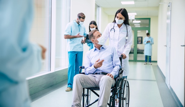 Больной старший мужчина с защитной маской на лице в инвалидной коляске и уверенный врач в медицинской маске во время транспортировки по больнице.
