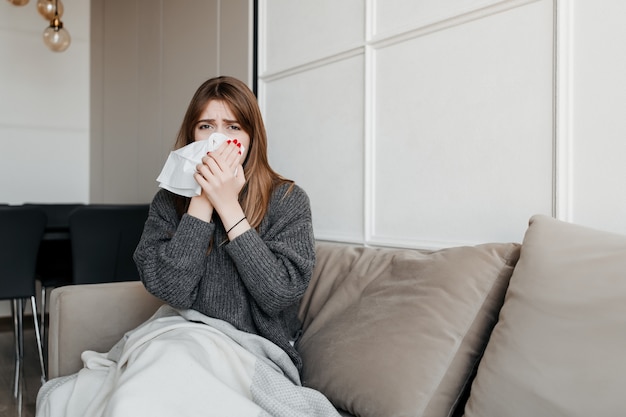 Ill woman blowing nose with tissue sitting on couch at home in apartment