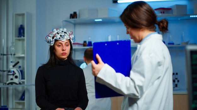 Ill woman listening neurological doctor looking at clipboard wearing high tech headset scanning brain electrical activity. Patient sitting in scientific laboratory treating nervous system dysfunctions