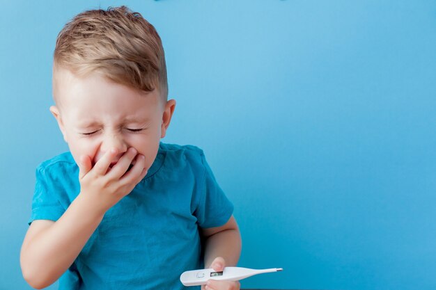 Ill young child with a thermomether, measuring the height of his fever and looking into the camera