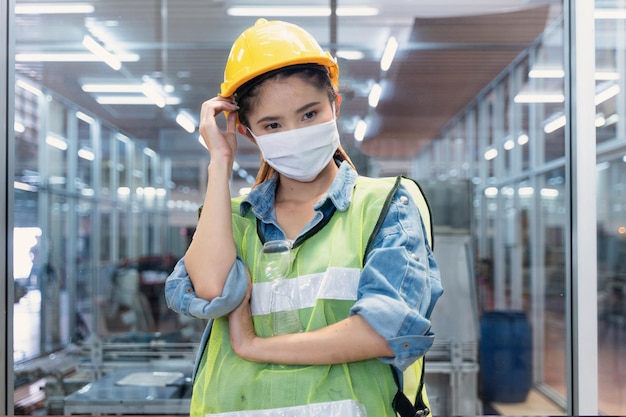 사진 마스크 덮개 얼굴을 가진 질병 제조 노동자 여성은 첨단 산업 공장의 유리 벽 앞에 서 있습니다. 스마트 산업 작업자 운영의 개념입니다.