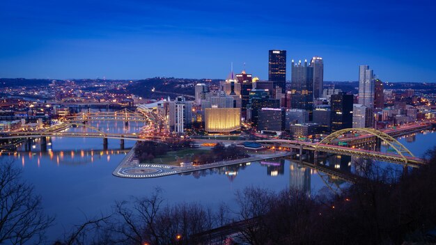 Illuminated bridge over river by buildings against sky at dusk