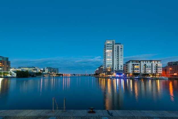 Illuminated buildings by river against blue sky at dusk