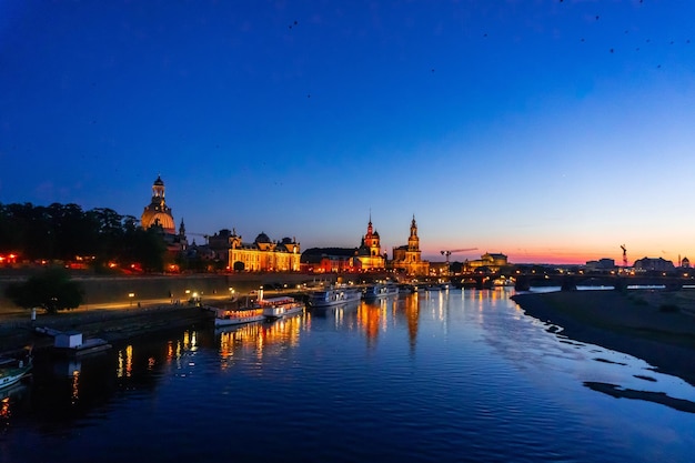 illuminated buildings by river against sky at dusk