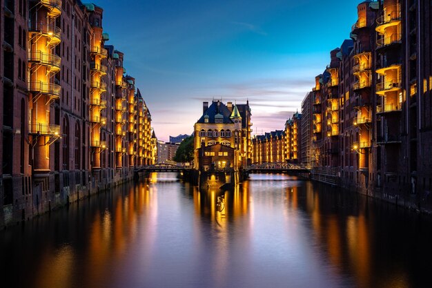 Illuminated buildings by river at sunset