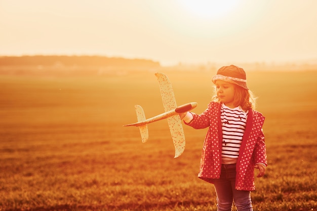 Illuminated by orange colored sunlight. Cute little girl have fun with toy plane on the beautiful field at daytime