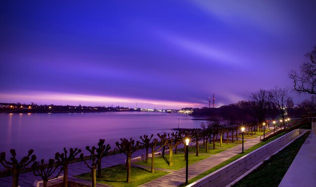 Illuminated city by river against sky at sunset