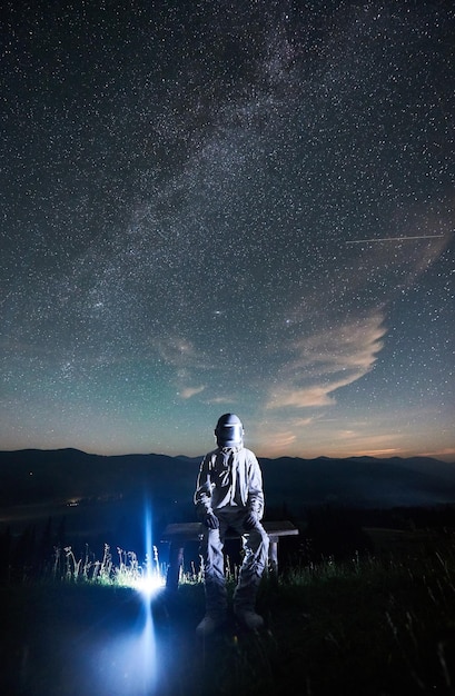 Illuminated man wearing white space suit and helmet sitting on a bench on a hill at night