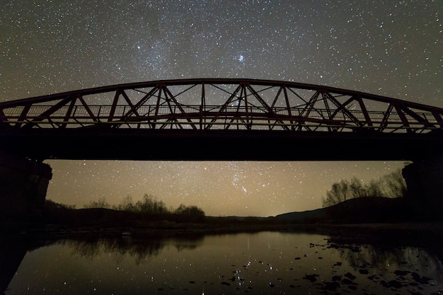 コンクリートのサポートに照らされた金属製の橋は、天の川星座の背景を持つ暗い星空の水に反映されます。夜の写真のコンセプト。