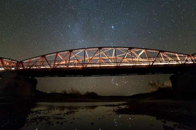 Illuminated metal bridge on concrete supports reflected in water on dark starry sky with Milky Way constellation. Night photography concept.