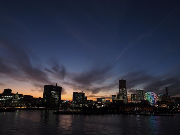 Illuminated modern buildings by lake against sky in city at dusk