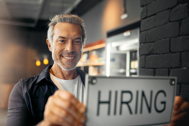 Im looking for some new staff members Cropped portrait of a handsome mature man holding up a hiring sign while standing in his coffee shop