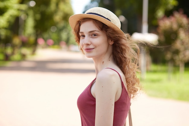 Image of adorable beautiful youngster looking aside, standing in middle of summer park, smiling sincerely, having pleasant look, wearing straw hat, red shirt, having bag on shoulder. Youth concept.