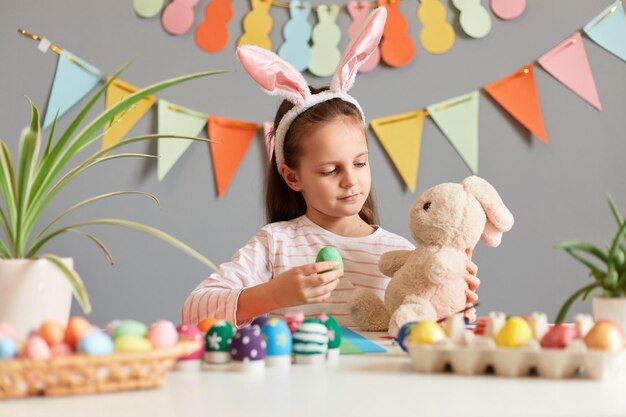 Изображение очаровательной милой маленькой девочки, одетой в кроличьи уши, сидящей за столом, держащей свою мягкую игрушку и показывающей свои красивые зеленые пасхальные яйца своему пушистому другу, позирующему на фоне украшенной серой стены