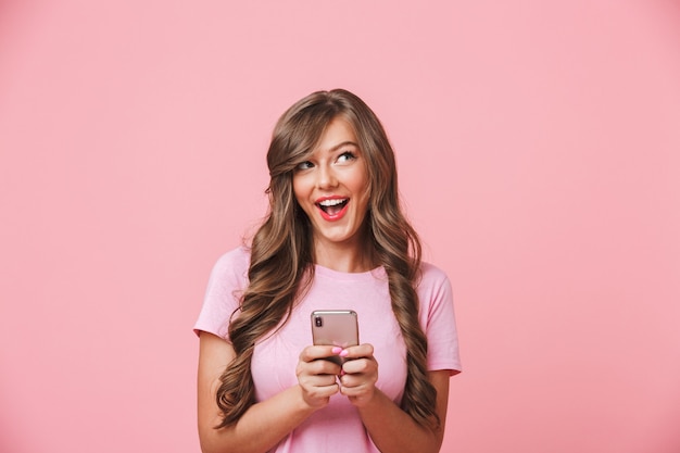 Image of adorable woman 20s with long curly hair and playful look holding mobile phone in hands while chatting, isolated over pink background