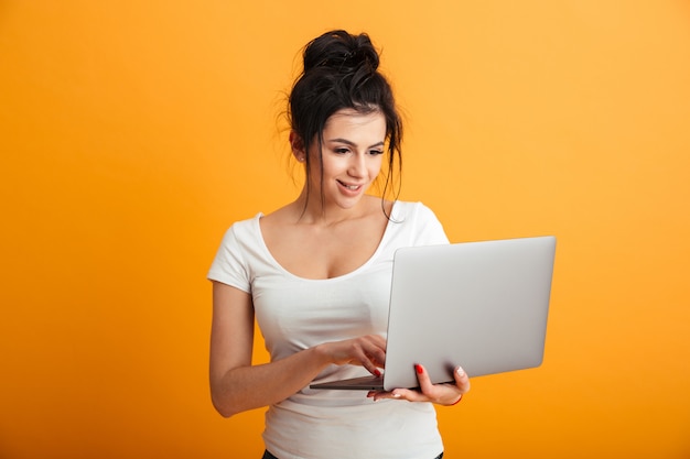 Image of adorable young woman in casual shirt looking on screen of notebook holding in hand, over yellow wall