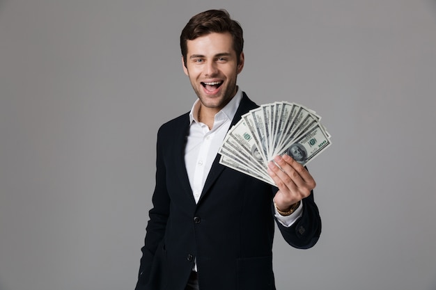 Image of adult businessman 30s in suit smiling and holding fan of money in dollar banknotes, isolated over gray wall