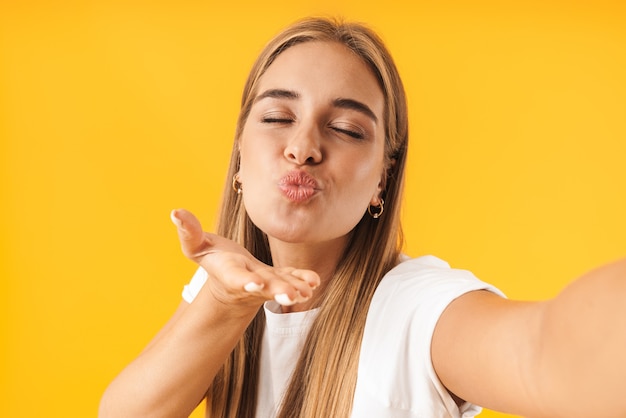 Photo image of alluring woman in basic clothes blowing air kiss  while taking selfie photo isolated over yellow wall