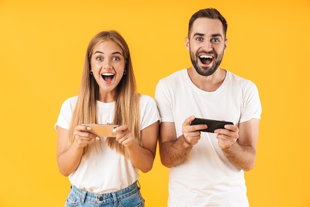 Image of amazed couple man and woman in basic t-shirts playing video games on smartphones together isolated over yellow wall