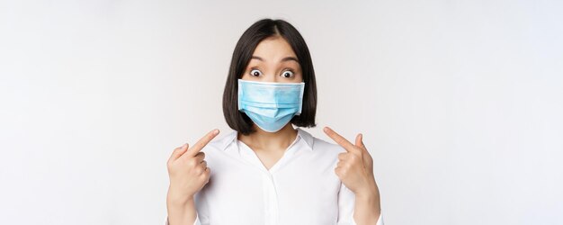 Image of amazed young asian woman pointing at herself while wearing medical face mask standing over white background