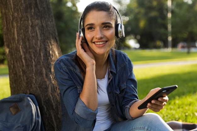 携帯電話を使用してヘッドフォンで音楽を聴いている公園の驚くべき美しい若い女性の学生の画像。