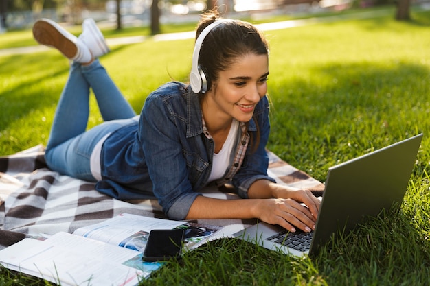 Photo image of amazing happy woman student lies outdoors in a park using laptop computer listening music.